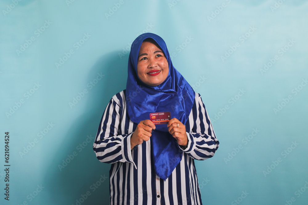 Excited Middle-aged Asian woman in blue hijab and striped shirt showing, presenting credit card for paying online business, isolated over blue background. Excited Middle-aged Asian woman in blue hijab and striped shirt showing, presenting credit card for paying online business, isolated over blue background.