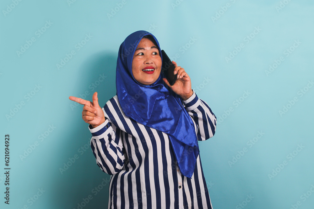 A middle-aged Asian woman in a blue hijab and a striped shirt is pointing to the left copyspace while talking on the phone. She is isolated on a blue background A middle-aged Asian woman in a blue hijab and a striped shirt is pointing to the left copyspace while talking on the phone. She is isolated on a blue background