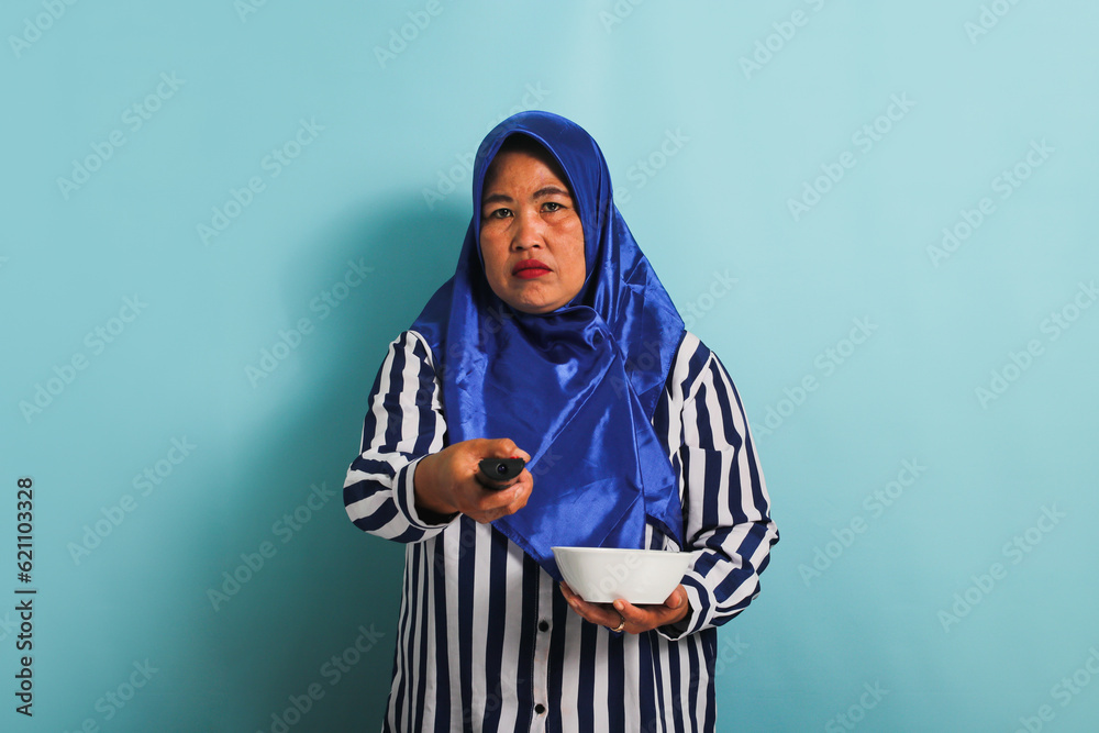 An unhappy middle-aged Asian woman in a blue hijab and a striped shirt is feeling bored while watching TV, switching channels with a TV remote, and holding a white bowl, isolated on a blue background. An unhappy middle-aged Asian woman in a blue hijab and a striped shirt is feeling bored while watching TV, switching channels with a TV remote, and holding a white bowl, isolated on a blue background.