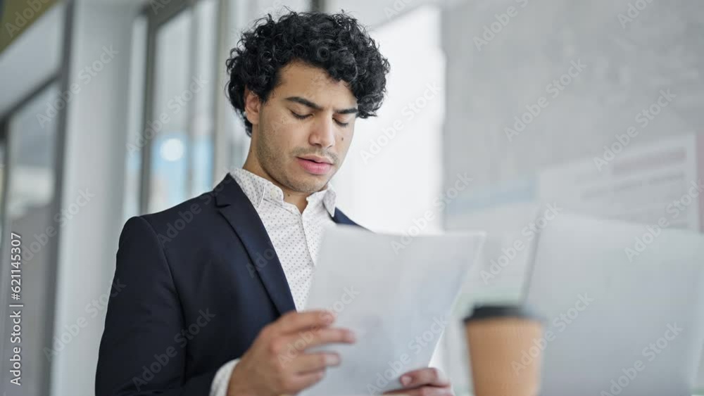 Young latin man business worker reading document sitting on table at office Young latin man business worker reading document sitting on table at office