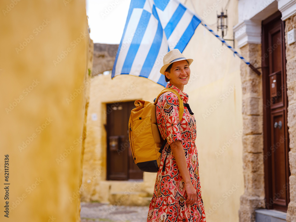 Young Asian woman in ethnic red dress walks and looks at cozy narrow streets of old city. Tourism, vacation, and discovery concept, female traveler visiting southern Europe, Rhodes island, Greece.