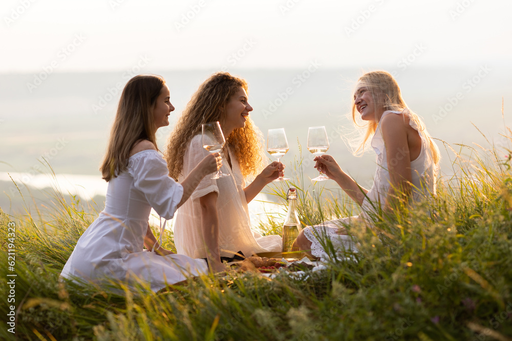 Three young happy girlfriends in elegant white dresses is having picnic on the hill on sunset.