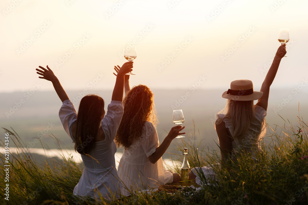 Beautiful girlfriends having picnic on the hill on summer sunset. They sitting with hands up and enjoying beautiful landscape.