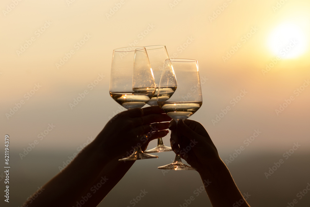 Close-up of hands holding glasses with white wine during sunset. Celebration and friendship concept.