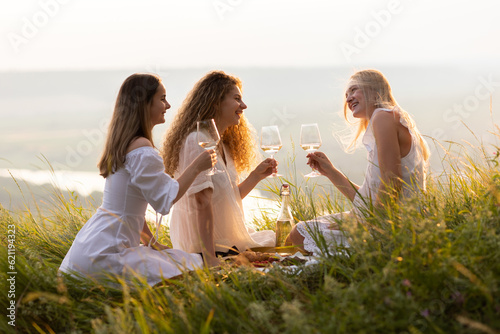 Obraz na płótnie Three young happy girlfriends in elegant white dresses is having picnic on the hill on sunset. w salonie