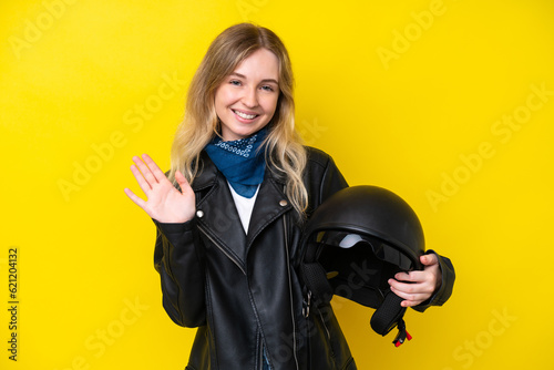 Blonde English young girl with a motorcycle helmet isolated on yellow background saluting with hand with happy expression