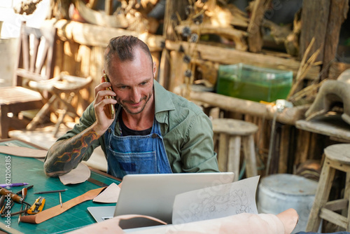 Closeup leather craftsman check and talking mobile with customer for orders in online shop with laptop at leather workplace.