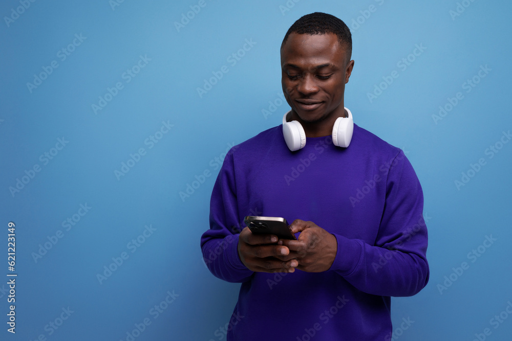 handsome young american man in blue sweatshirt with wireless bluetooth headphones and smartphone on blue background with copy space handsome young american man in blue sweatshirt with wireless bluetooth headphones and smartphone on blue background with copy space