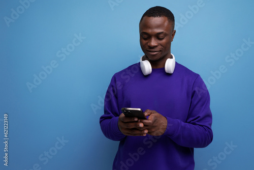 handsome young american man in blue sweatshirt with wireless bluetooth headphones and smartphone on blue background with copy space © Ivan Traimak