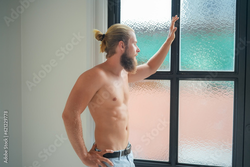 A young man is standing in bathroom with a large bathtub, he looks out the window.