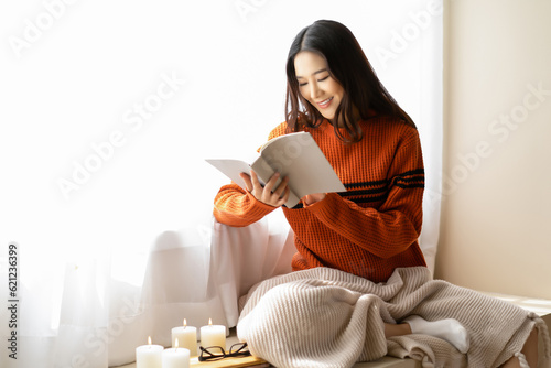 Beautiful young Asian woman reading book wearing knitted warm sweater sit on windowsill in room decorated with candle at home. Korea or Japanese girl relaxing at home. Cozy lifestyle, hygge concept