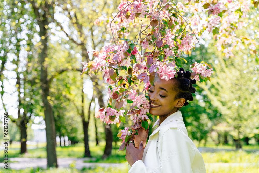 Obraz premium Concept of relaxation and mental health. African american teenager girl with flowering branch of sakura tree in spring blooming garden enjoying warm sunny weather.