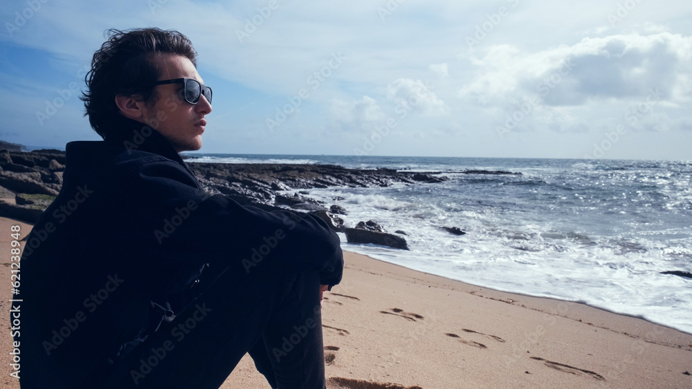 Thoughtful handsome young man sits on the empty sandy beach on the summer day. Guy is sitting on sandy beach on a sunny day. Man with a romantic mood on the beach contemplating the beautiful ocean Thoughtful handsome young man sits on the empty sandy beach on the summer day. Guy is sitting on sandy beach on a sunny day. Man with a romantic mood on the beach contemplating the beautiful ocean