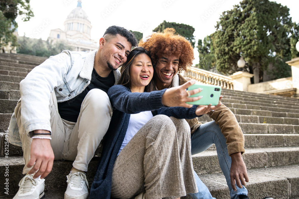 Three young diverse people taking a selfie with a smartphone sitting on a stairs. Multiracial group laughing and taking a picture with a phone in the park.