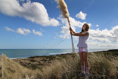 Happy girl age 11 above remote scenic New Zealand beach, holding native toetoe fronds. Surfers in the distance.