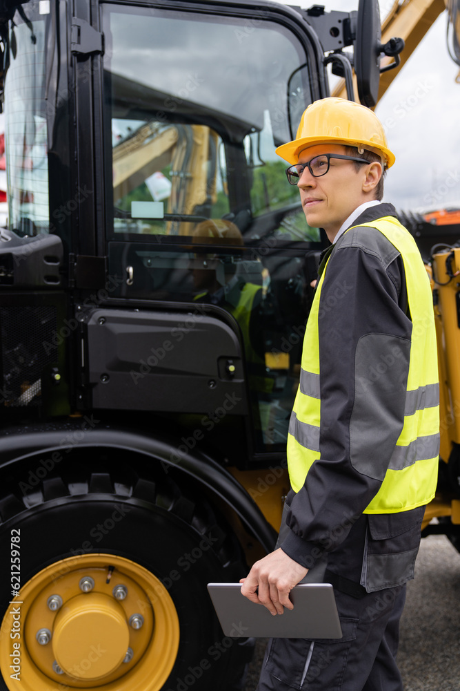 Engineer in a helmet with a digital tablet next to construction excavators Engineer in a helmet with a digital tablet next to construction excavators