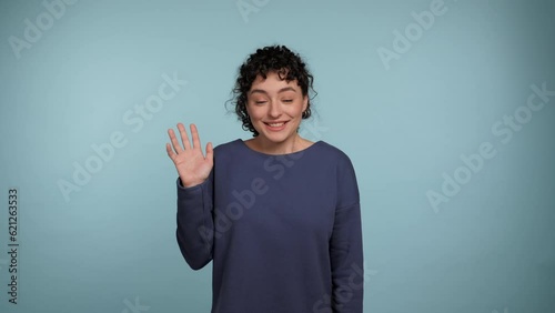 Smiling young curly woman greets by waving her hand, showing hello gesture looks into camera. Portrait positive female standing on isolated light blue background. People emotions lifestyle concept