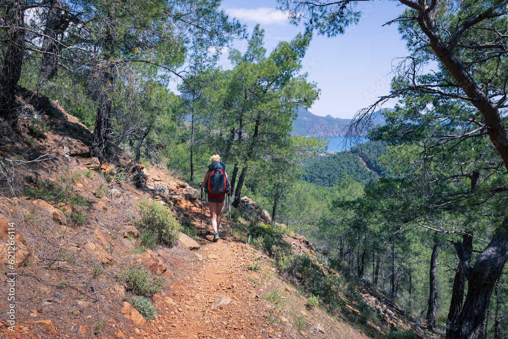 Obraz premium Woman with backpack and trekking poles walks on marked path on forested hillside, Lycian Way, Turkey