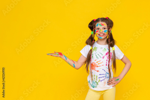 A child smeared with paint. A little girl, painted with multicolored paints, holds your advertisement in the palm of her hand. Children's creativity. Yellow isolated background. Copy space. Banner.
