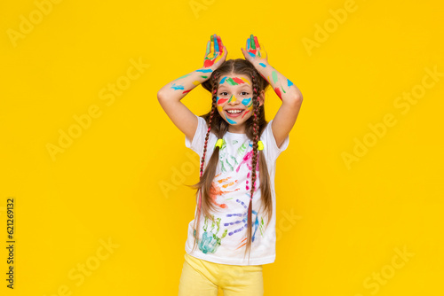 Portrait of a girl with a face painted with multicolored paints. Children's creativity. The child shows his ears above his head. Yellow isolated background.