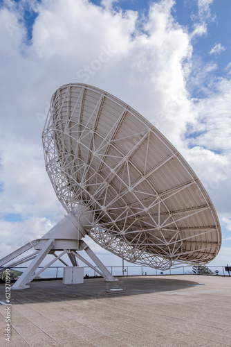 Side view of large satellite dish to observe the cosmos, with large white clouds in the background.