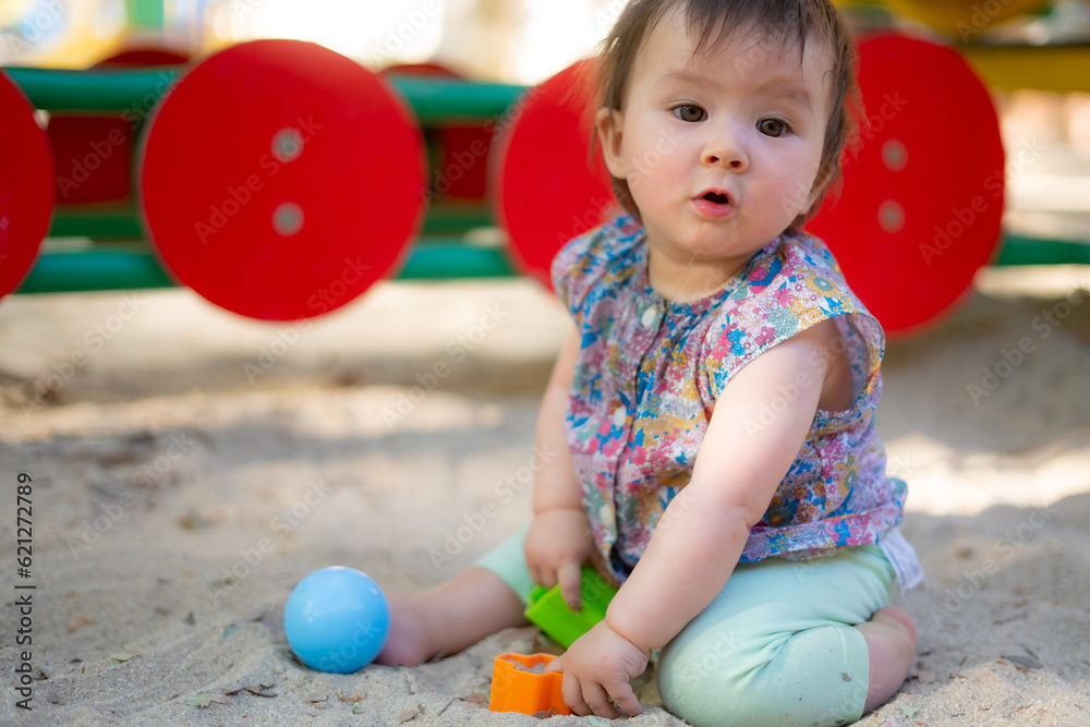 Obraz premium Adorable 9 months old baby playing outdoors - lifestyle portrait of mixed ethnicity Asian Caucasian baby girl playing with block toys happy and carefree at playground sitting on sand