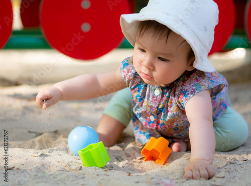 Adorable 9 months old baby playing outdoors - lifestyle portrait of mixed ethnicity Asian Caucasian baby girl playing with block toys happy and carefree at playground sitting on sand