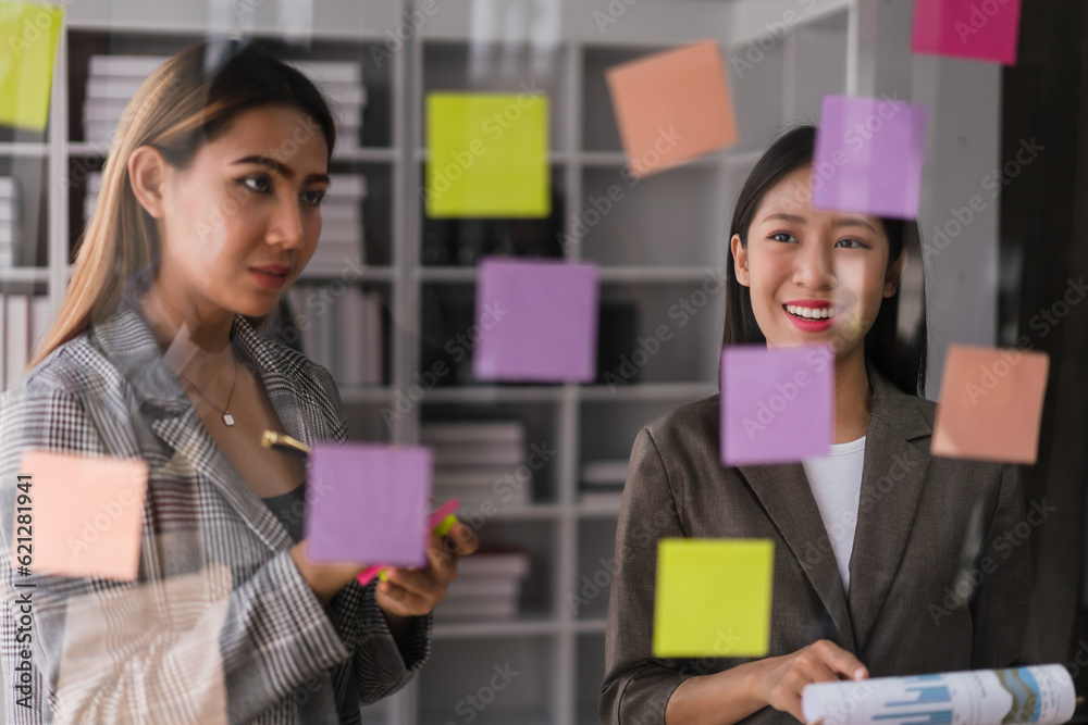 Two businesswomen brainstorming strategy of new business project and writing ideas on sticky notes Two businesswomen brainstorming strategy of new business project and writing ideas on sticky notes