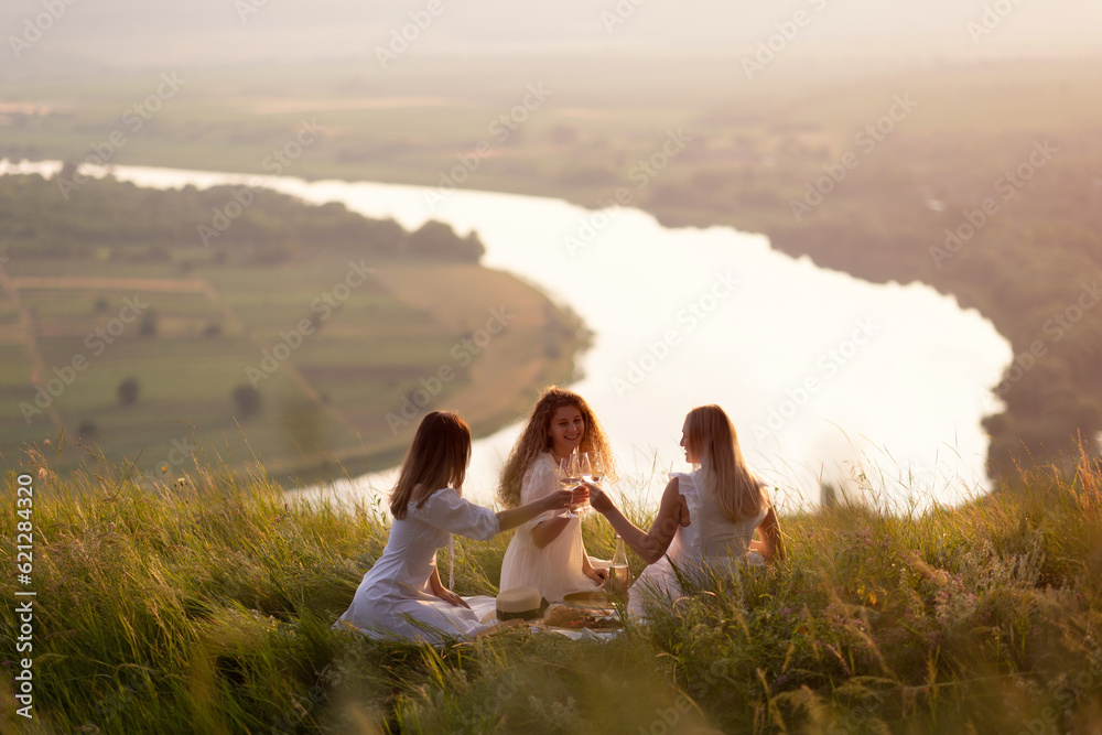 Young girlfriends drinking wine, laughing, having fun together at picnic at summer sunset. Slumber party. Summer picnic party ideas. Outdoor gathering with friends.