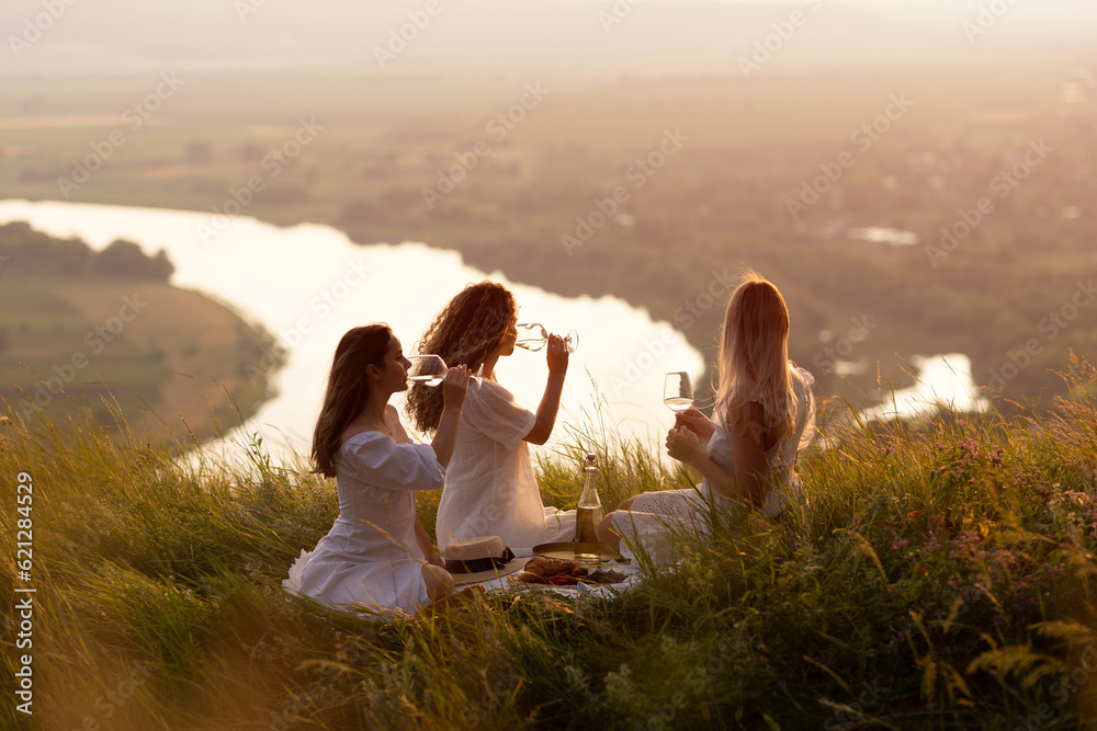 Friends having picnic on the hill at sunset. Three girlfriends eating and drinking wine on outdoor party at sunset.