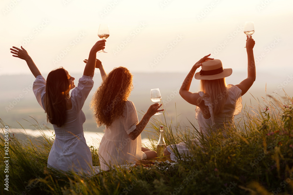 Beautiful girlfriends having picnic on the hill on summer sunset. They sitting with hands up and enjoying beautiful landscape.