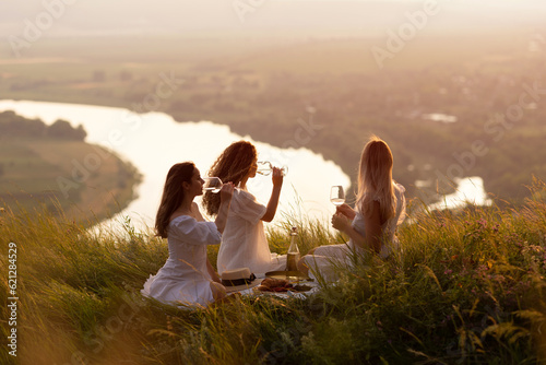 Friends having picnic on the hill at sunset. Three girlfriends eating and drinking wine on outdoor party at sunset.