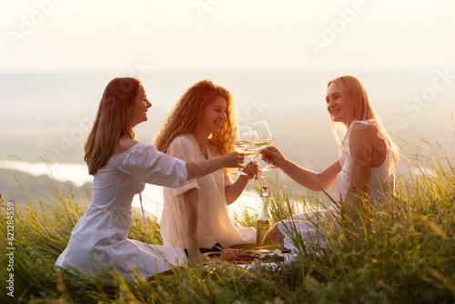 Obraz na płótnie Three young beautiful smiling female in summer white dresses. Carefree women making picnic in the park at sunset. Positive models sitting on plaid on grass, drinking wine, eating fruits and cheese. w salonie