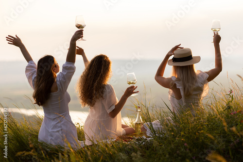 Obraz na płótnie Back view of girlfriends in stylish summer clothes raising arms with wine at sunset during picnic in summer park-
w salonie