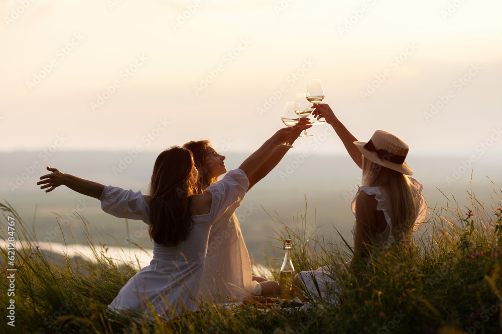 Company of young female friends having fun, raising glasses with wine and enjoy beautiful sunset at summer picnic on the hill.