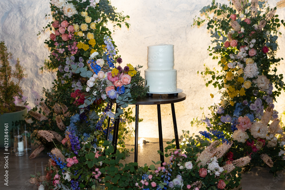 White wedding cake in the evening. Cake on the background of an arch of fresh flowers.