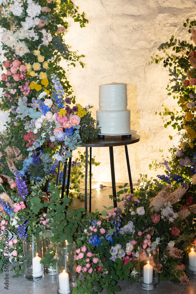 White wedding cake on a background of a flower arch. Wedding decor with many fresh flowers.