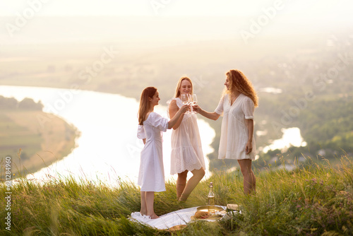 Obraz na płótnie Young happy girlfriends having fun at picnic during sunset. Food and beverage concept at nature. w salonie