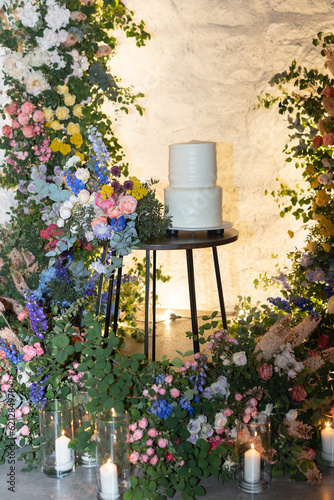 White wedding cake on a background of a flower arch. Wedding decor with many fresh flowers.	