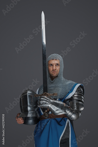 Proud knight stands holding his sword and helmet, donning blue surcoat, chainmail coif, and medieval armor, against a gray backdrop