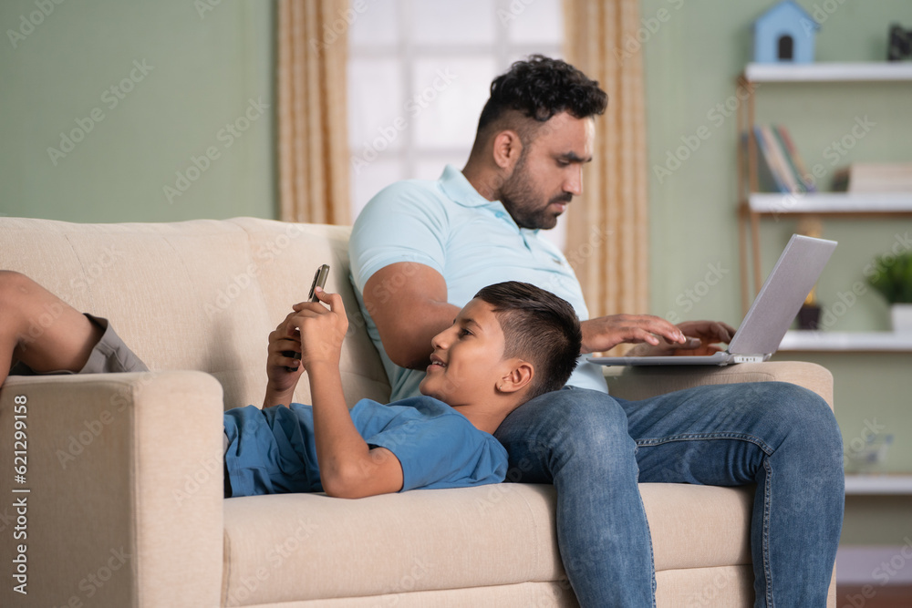 Indian father working on laptop while son using mobile phone by sleeping on lap on sofa at home - concept of Digital connection, technology addiction and cyberspace. Indian father working on laptop while son using mobile phone by sleeping on lap on sofa at home - concept of Digital connection, technology addiction and cyberspace.