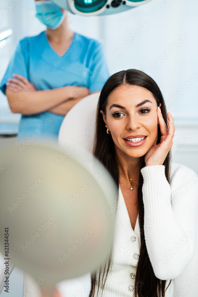 Fototapeta premium Beautiful and happy young woman sitting in medical chair and looking in the mirror. She is satisfied after successful beauty treatment with hyaluronic acid fillers or botulinum toxin injections.
