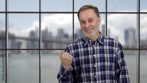 Portrait of smiling mature man pointing back with his thumb up. Window with city river view in the background. photo