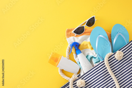 Skincare rituals during a summer holiday. Top view flat lay of sunscreen bottles, beach bag, striped towel, flip-flops, eyewear on yellow background with empty space for promo or text