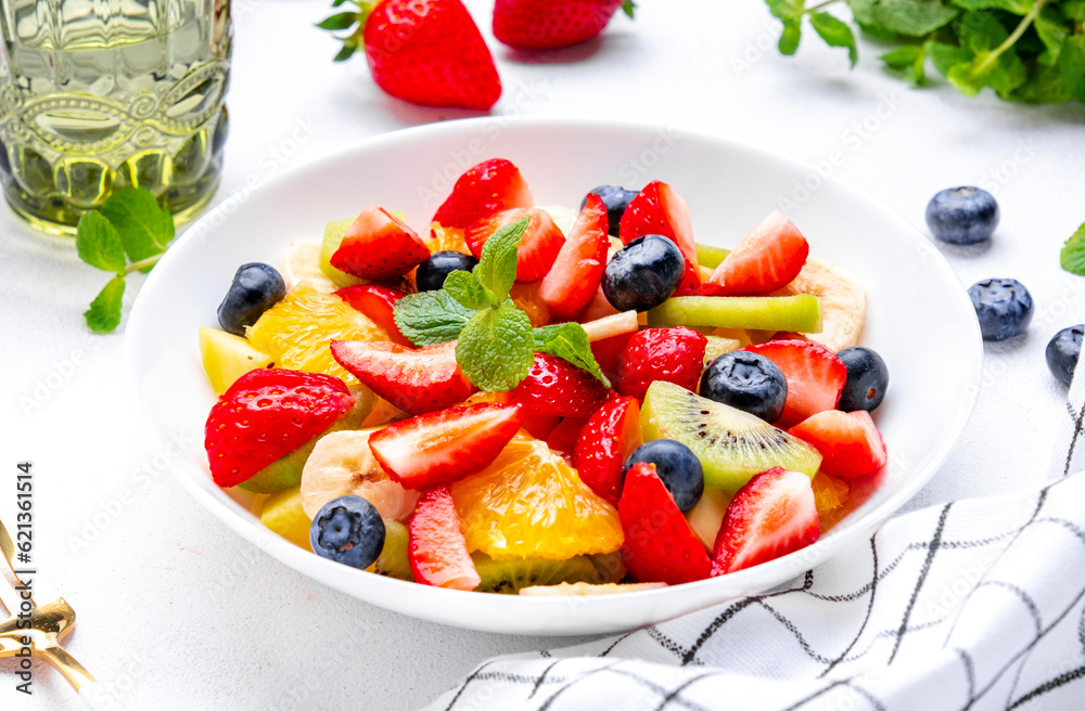 Fruit and berry salad with fresh strawberries, blueberries, banana, kiwi, orange and mint leaves, white table background, top view