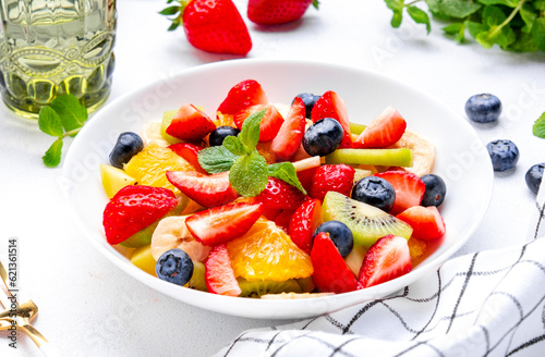 Fruit and berry salad with fresh strawberries  blueberries  banana  kiwi  orange and mint leaves  white table background  top view