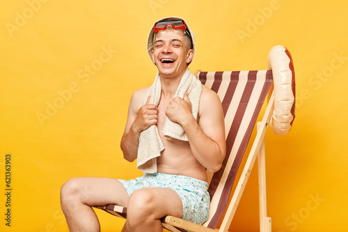 Attractive laughing shirtless man sitting on deck chair isolated over yellow background enjoying time at resort having fun holding towel on shoulders.