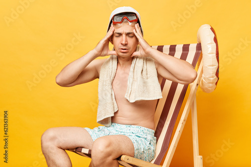 Sick shirtless man sitting on deck chair isolated over yellow background spending lots hours sunbathing suffering headache has insolation. photo