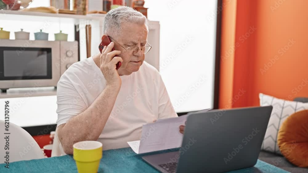 Middle age grey-haired man talking on smartphone reading document at dinning room Middle age grey-haired man talking on smartphone reading document at dinning room