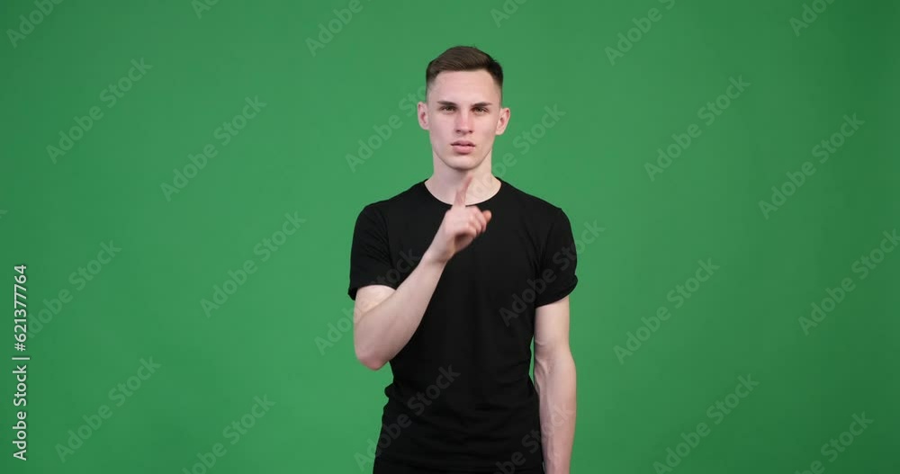 On a green background, a serious Caucasian man raises his index finger to his lips, signaling the shush or hush gesture. With a focused expression, he presses his finger against his lips. On a green background, a serious Caucasian man raises his index finger to his lips, signaling the shush or hush gesture. With a focused expression, he presses his finger against his lips.